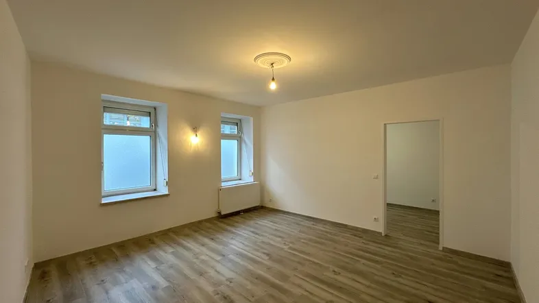 Helles, leeres Zimmer mit Laminatboden und zwei Fenstern, bereit zur individuellen Gestaltung.