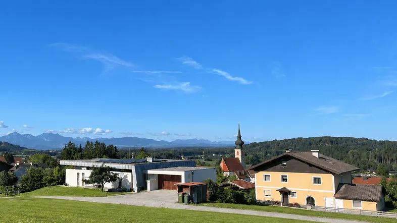 Panoramablick auf eine ländliche Siedlung mit Kirche und Alpen im Hintergrund unter blauem Himmel.