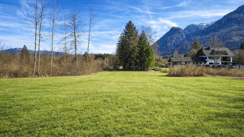 Weitläufige Grünfläche mit Blick auf ein Wohnhaus und die umliegende Berglandschaft unter blauem Himmel.