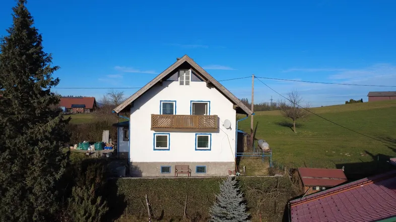Traditionelles Haus mit Balkon und Satteldach, umgeben von grünen Hügeln unter blauem Himmel.