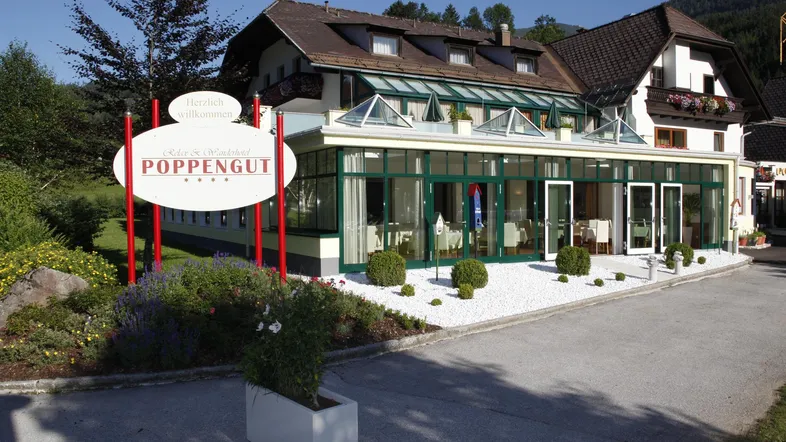 Eingangsbereich eines Hotels mit Wintergarten-Restaurant und gepflegter Außenanlage unter blauem Himmel.