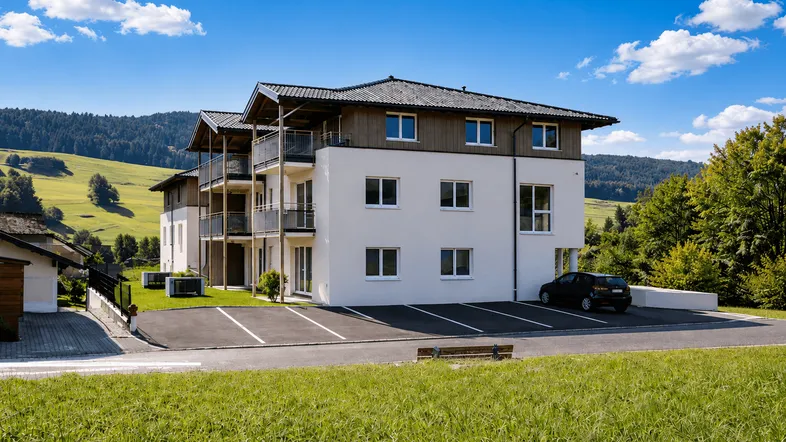 Modernes Mehrfamilienhaus mit Balkonen und Parkplätzen, eingebettet in eine grüne Hügellandschaft.