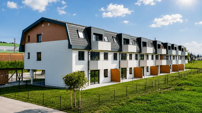 Moderne Reihenhäuser mit Satteldach und Balkonen, umgeben von grünen Wiesen unter blauem Himmel.