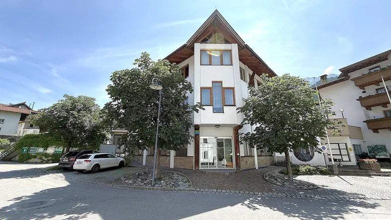 Modernes Gebäude mit spitzem Dach und großen Fenstern, umgeben von Bäumen und Parkplätzen unter blauem Himmel.