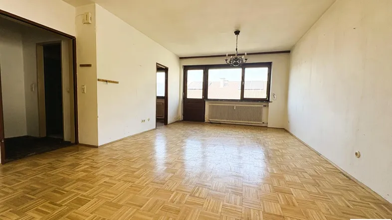 Helles, leeres Wohnzimmer mit Fischgrätparkett und einem Fenster, das Tageslicht hereinlässt.