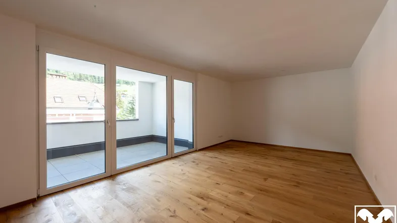 Helles, leeres Wohnzimmer mit Holzboden und Zugang zu einem Balkon.