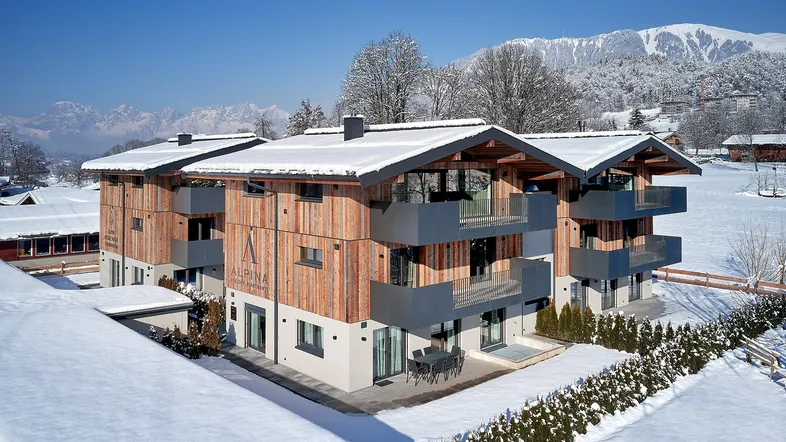 Modernes Apartmentgebäude in den Alpen, umgeben von Schnee und Bergen unter blauem Himmel.