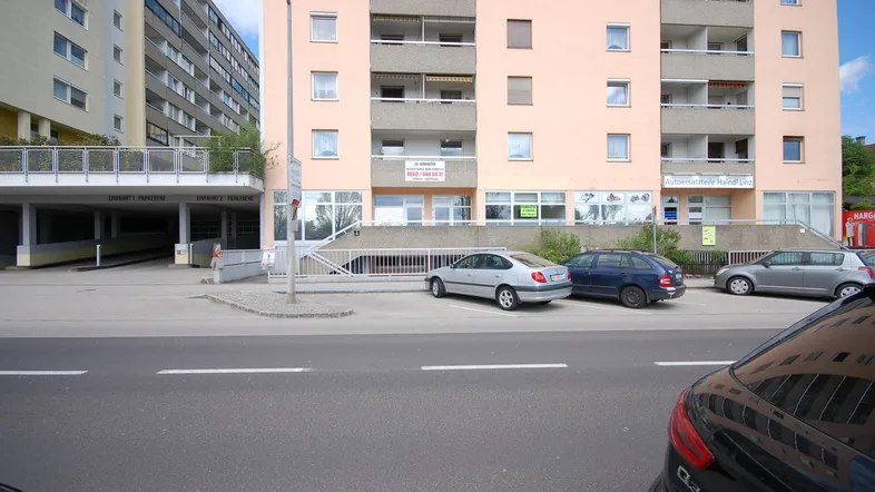 Mehrstöckiges Wohngebäude mit Geschäften im Erdgeschoss und Parkplätzen davor an der Straße.