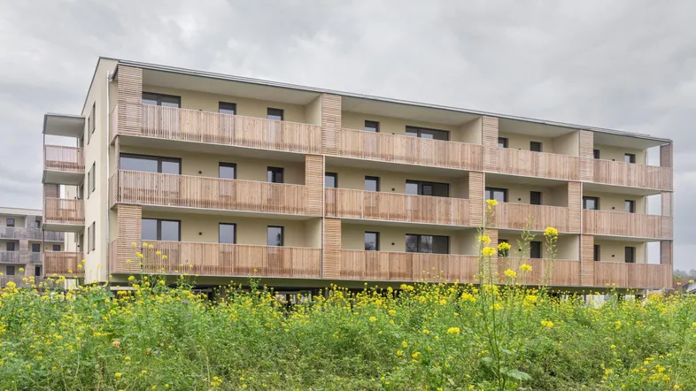 Modernes Mehrfamilienhaus mit Holzfassade und Balkonen, umgeben von blühenden Feldern unter bewölktem Himmel.