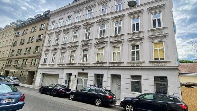 Klassische Fassade eines Altbaus mit mehreren Etagen und parkenden Autos auf der Straße unter bewölktem Himmel.