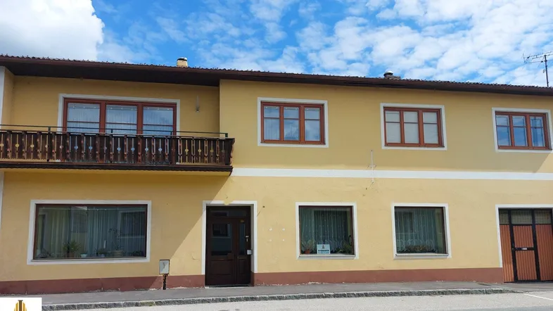 Gelbe Fassade eines Mehrfamilienhauses mit Holzbalkon und braunen Fensterrahmen unter blauem Himmel.