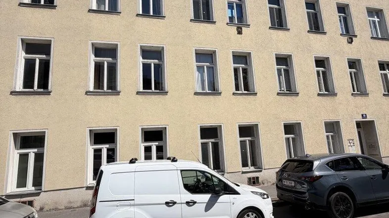 Fassade eines Mehrfamilienhauses mit mehreren Fenstern und geparkten Autos davor.