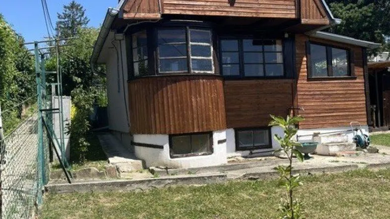 Rustikales Holzhaus mit Mansarddach und Gartenbereich unter blauem Himmel.