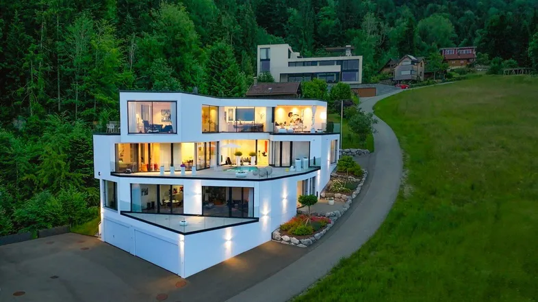 Moderne Villa mit markanter Architektur, großen Glasfronten und beleuchteten Terrassen in grüner Hanglage.
