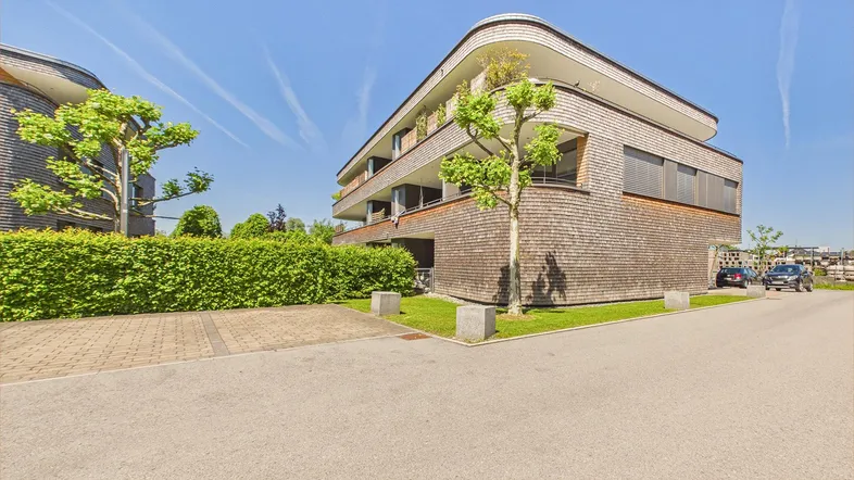 Modernes Gebäude mit Holzfassade, umgeben von Hecken und Grünflächen unter blauem Himmel.