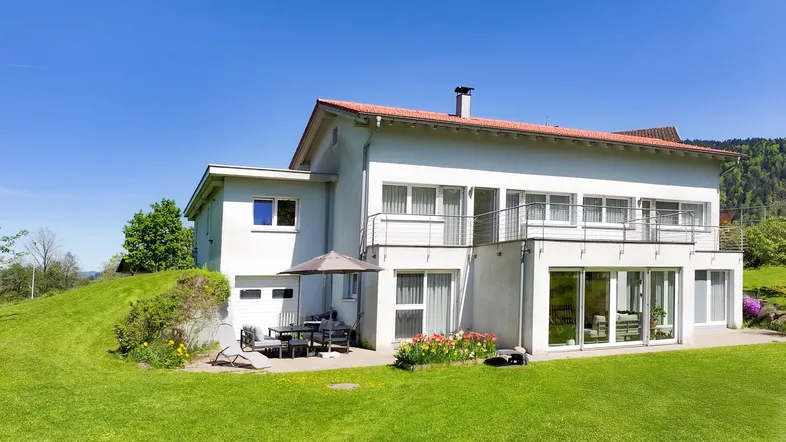 Weißes Einfamilienhaus mit rotem Dach, großem Garten und möblierter Terrasse unter blauem Himmel.