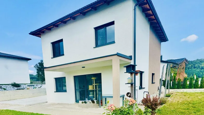 Modernes Einfamilienhaus mit weißer Fassade, dunklen Fenstern und einem gepflegten Vorgarten unter blauem Himmel.