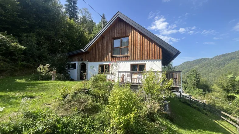 Charmantes Haus mit Holzfassade und kleiner Terrasse, eingebettet in grüne Hügellandschaft.