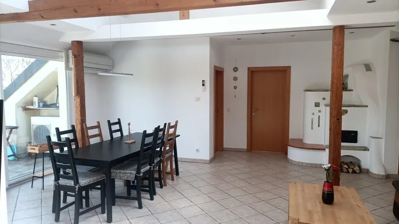 Geräumiger Wohn-Essbereich mit Holzbalken, Kachelofen und Zugang zu einem Außenbereich.