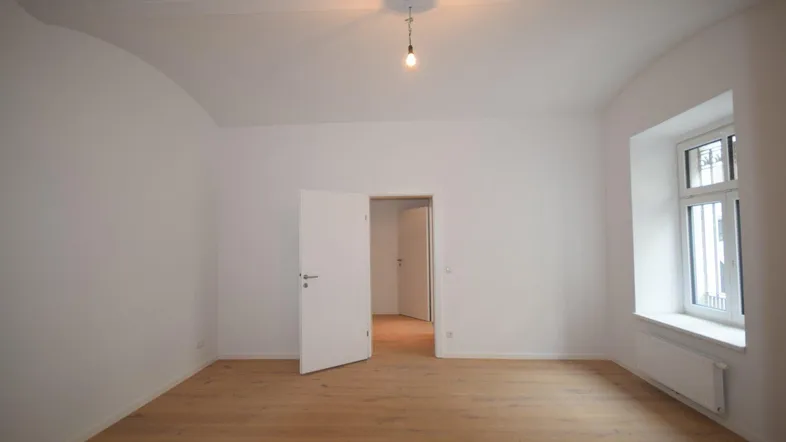 Helles, leeres Zimmer mit hohen Decken, Holzboden und einem Fenster, das viel Tageslicht hereinlässt.