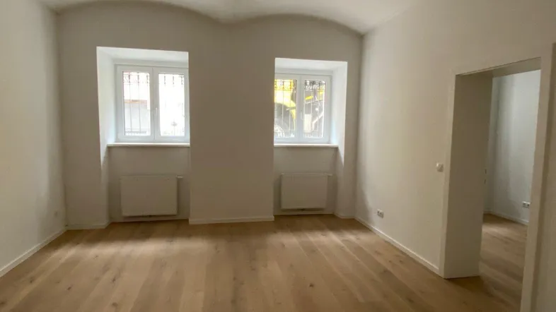 Heller Raum mit Holzboden, weißen Wänden und zwei Fenstern, ideal als Wohnzimmer.