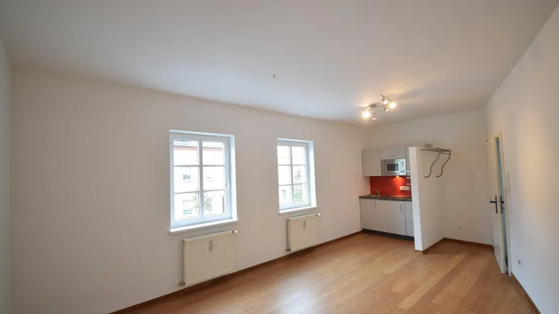 Helles Wohnzimmer mit Holzboden, zwei Fenstern und integrierter Küchenzeile.