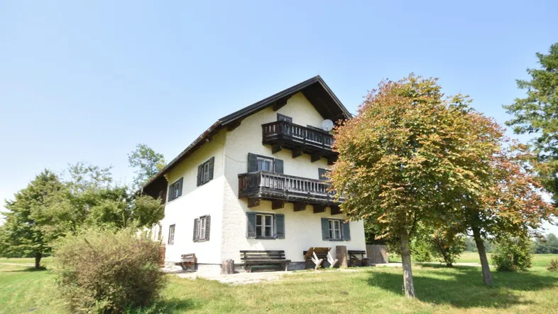 Traditionelles Haus mit weißer Fassade, Holzbalkonen und Satteldach, umgeben von grüner Landschaft unter blauem Himmel.