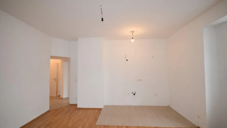 Heller Raum mit Holzboden und vorbereiteten Anschlüssen für eine zukünftige Küche.