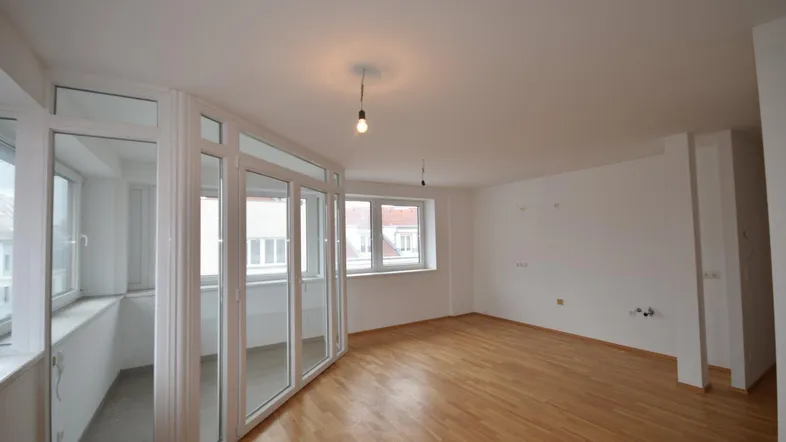 Helles, leeres Wohnzimmer mit Parkettboden und einem verglasten Balkonbereich.