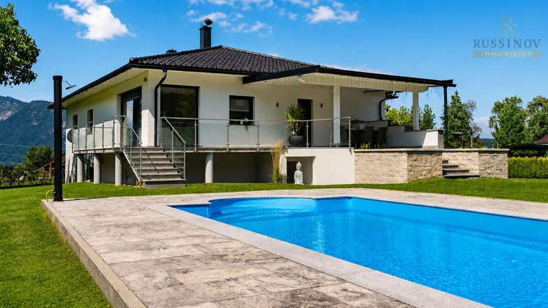 Modernes Haus mit großem Pool, weitläufigem Garten und Bergblick unter blauem Himmel.