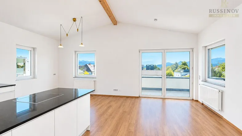Heller offener Wohn-Essbereich mit moderner Küche, Holzbalken und Zugang zur Terrasse mit Bergblick.