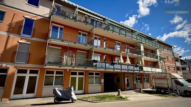 Mehrfamilienhaus mit mehreren Balkonen und Geschäften im Erdgeschoss unter blauem Himmel.