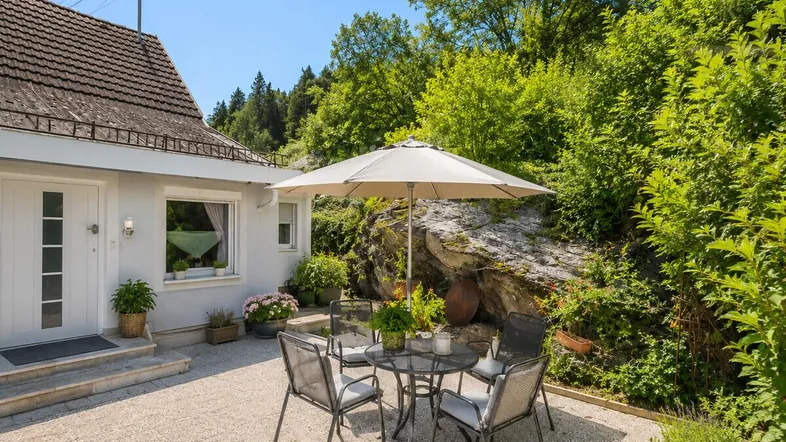 Gemütliche Terrasse mit Gartenmöbeln und Sonnenschirm, umgeben von grüner Natur und Felsen.