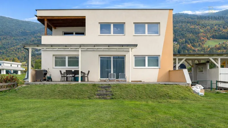 Modernes Haus mit Terrasse und Balkon, umgeben von grüner Natur und Bergen im Hintergrund.