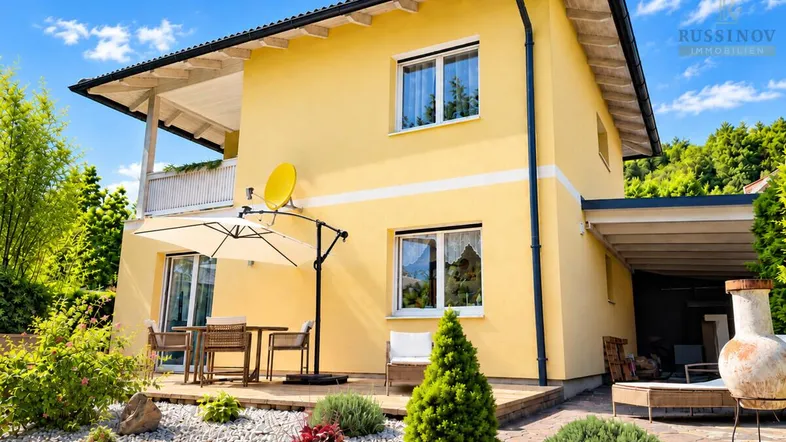 Gelbes Einfamilienhaus mit Terrasse, Gartenbereich und Bergblick unter blauem Himmel.