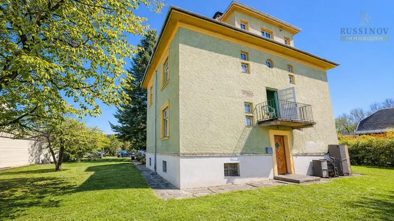 Historisches Gebäude mit hellgrüner Fassade, gelben Akzenten und kleinem Balkon in ländlicher Umgebung.