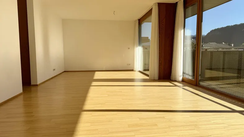 Helles, leeres Zimmer mit Holzboden und großen Fenstern, die viel Sonnenlicht hereinlassen.