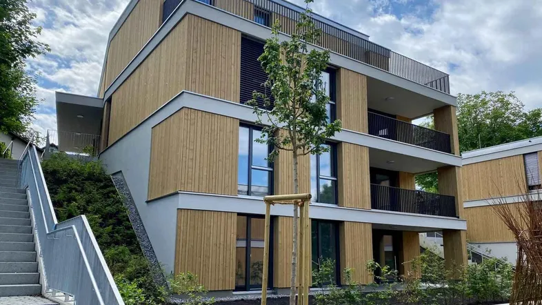 Modernes Mehrfamilienhaus mit Holzfassade, Balkonen und Außentreppe unter bewölktem Himmel.