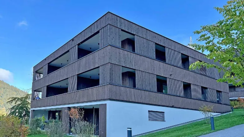 Modernes Mehrfamilienhaus mit Holzfassade und Balkonen, eingebettet in eine grüne Umgebung.