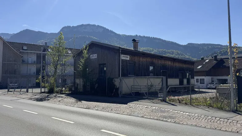 Gebäude mit Holzfassade an einer Straße, umgeben von Bäumen und Bergen im Hintergrund.