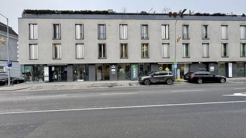 Modernes Geschäftsgebäude mit mehreren Ladenlokalen im Erdgeschoss und Wohnungen darüber, an einer Straße gelegen.
