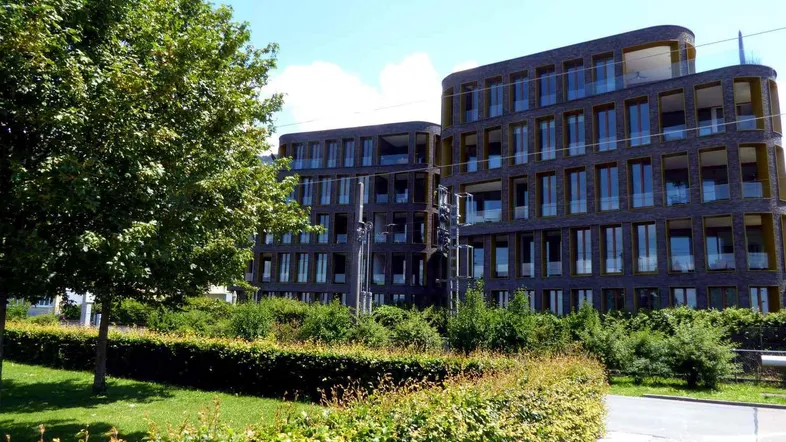 Moderne Wohngebäude mit dunkler Ziegelfassade und großen Fenstern, umgeben von grünen Hecken und Bäumen.