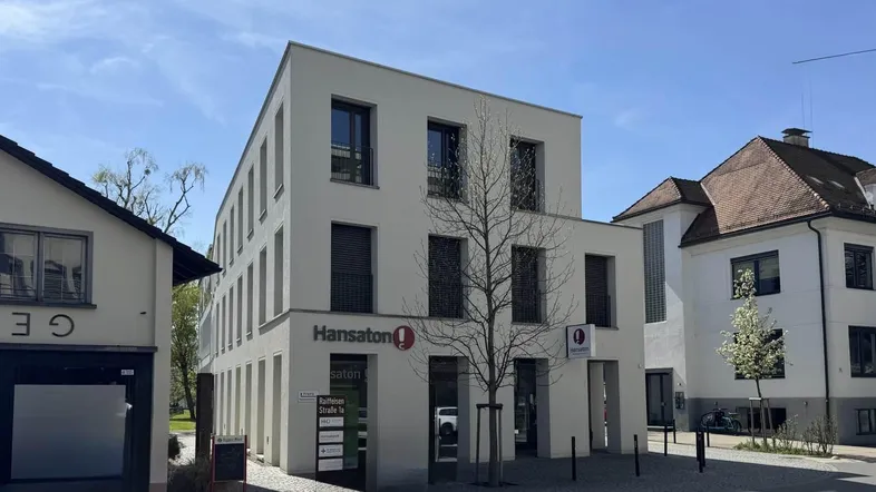 Modernes Geschäftsgebäude mit heller Fassade und großen Fenstern an einer städtischen Straße, umgeben von Bäumen.
