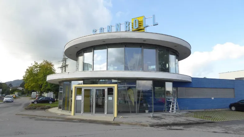 Modernes, rundes Gebäude mit großer Glasfassade und umliegenden Parkplätzen unter bewölktem Himmel.