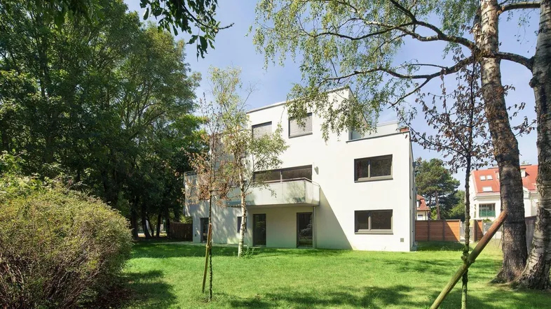 Modernes Wohngebäude mit weißer Fassade, umgeben von einem grünen Rasen und Bäumen unter blauem Himmel.