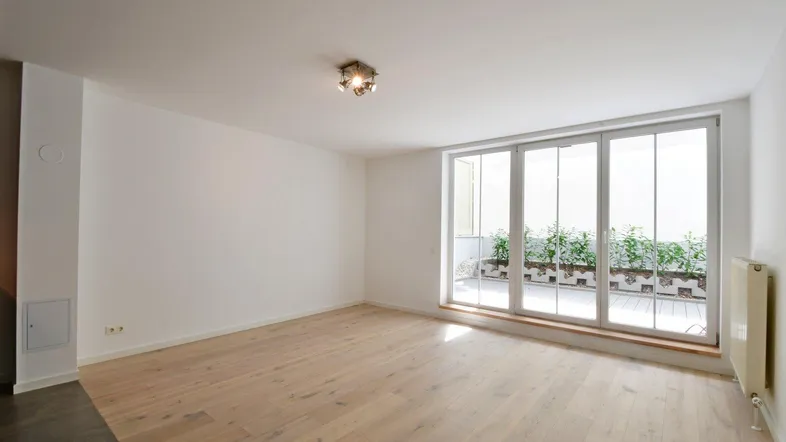 Helles, leeres Zimmer mit Holzboden und großen Fenstern, die zu einer Terrasse führen.