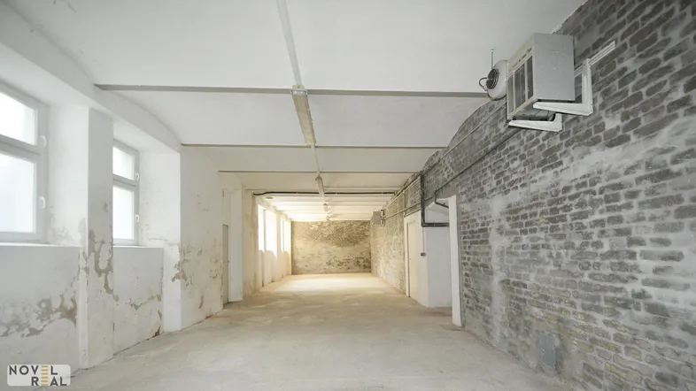 Langer, leerer Raum mit unverputzten Ziegelwänden, Betonboden und sichtbaren Lüftungsanlagen.