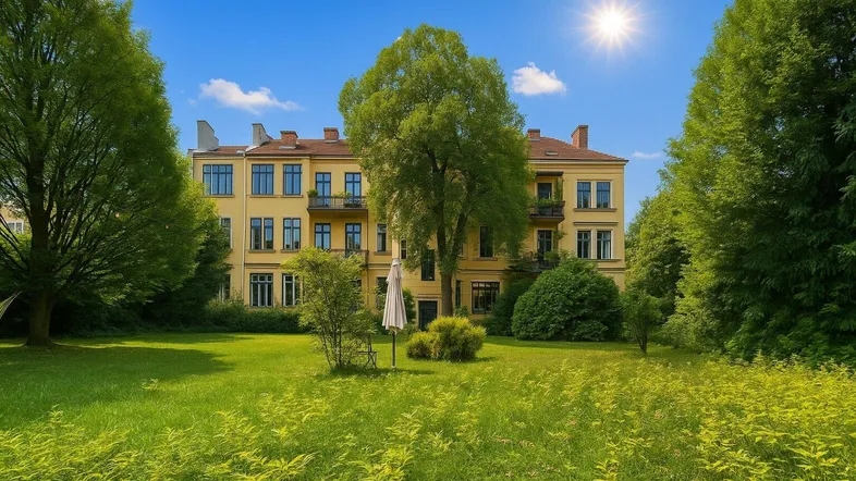 Gelbes Altbaugebäude mit vielen Fenstern und einem weitläufigen grünen Garten unter blauem Himmel.