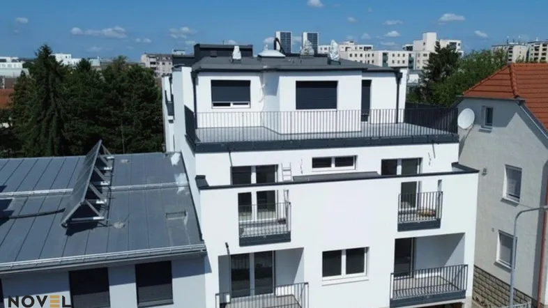 Modernes Mehrfamilienhaus mit weißer Fassade, Balkonen und einer Dachterrasse in städtischer Umgebung.