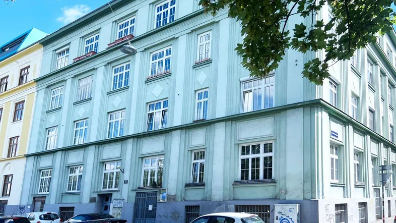 Grünes Mehrfamilienhaus mit vielen Fenstern und parkenden Autos auf der Straße.
